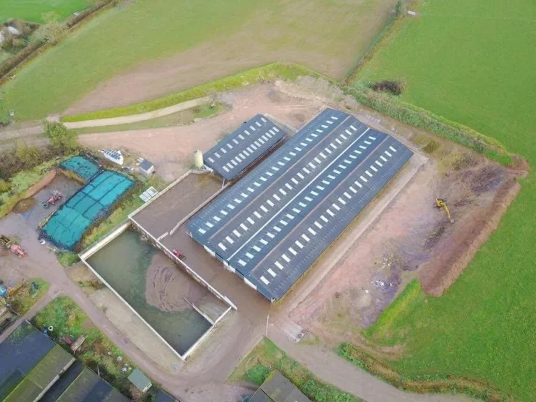 A spacious, dark green storage facility with a metal roof, nestled in a countryside landscape of gently rolling fields. Prominent sliding doors and a nearby digger add to its utilitarian charm.