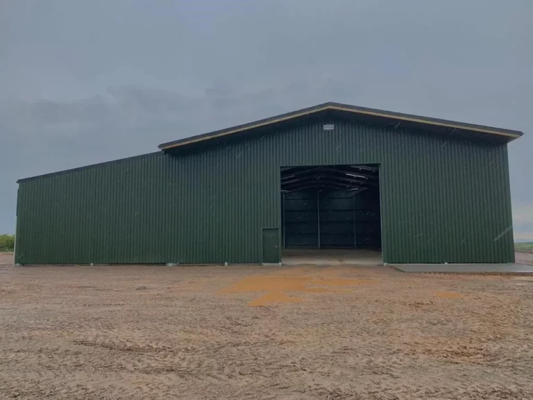 This image depicts a large, green agricultural steel building with a gabled roof. The structure is situated on a spacious, open, and unpaved ground, suggesting a rural or farm setting. The building features a wide entrance with a sliding door and an adjacent smaller pedestrian door. The design and construction indicate it is likely used for storage, such as for equipment, machinery, or harvested crops. The building's robust steel framework provides durability and protection against various weather conditions. This type of steel building is often chosen for its cost-effectiveness, ease of assembly, and low maintenance requirements, making it an ideal solution for agricultural storage needs.